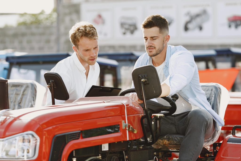 Two men discussing and evaluating the features of a red tractor outdoors in a dealership setting.